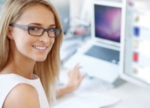 Mujer rubia con gafas de pasta sonriendo hacia la cámara trabajando en dos ordenadores